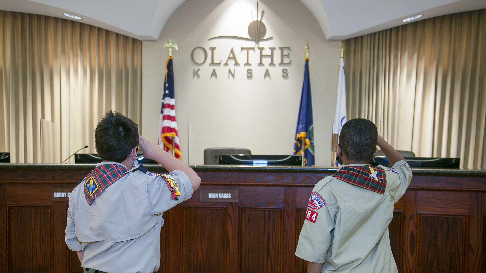 Two boy scouts stand with their backs to the camera and salute the Kansas and United States flag in the Olathe council chambers.