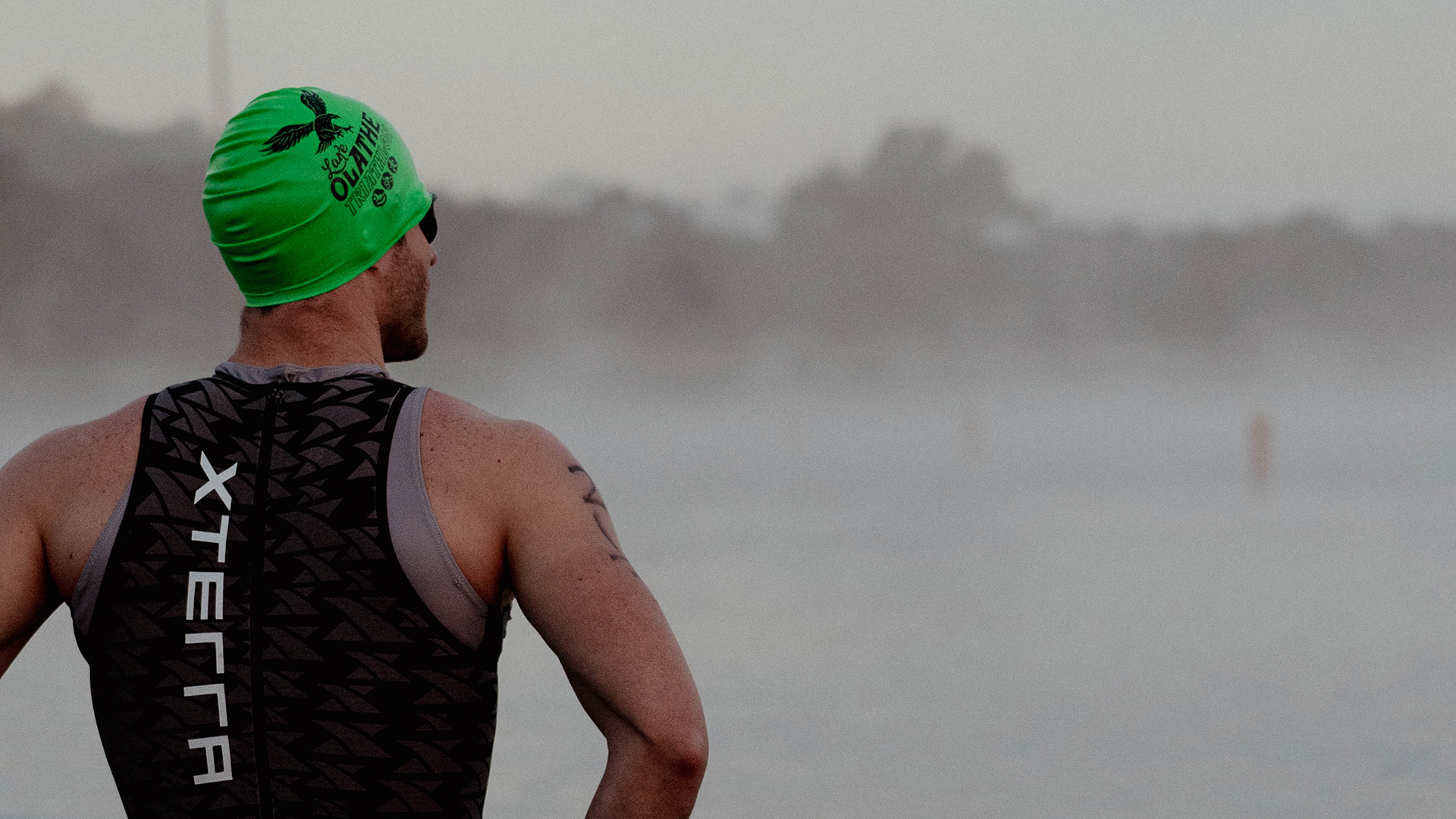 A Lake Olathe Triathlon athlete stands facing the lake, wearing a green swim cap with the Lake Olathe Triathlon logo