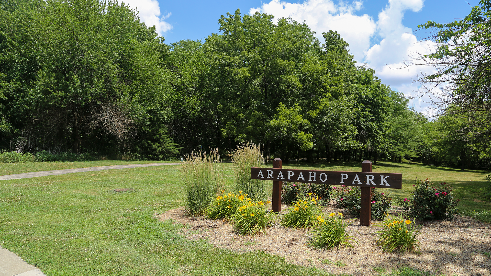 Arapaho Park in Olathe. Green space with a mulched landscape bed with green grassy bushes and a brown wooden sign that reads "Arapaho Park"