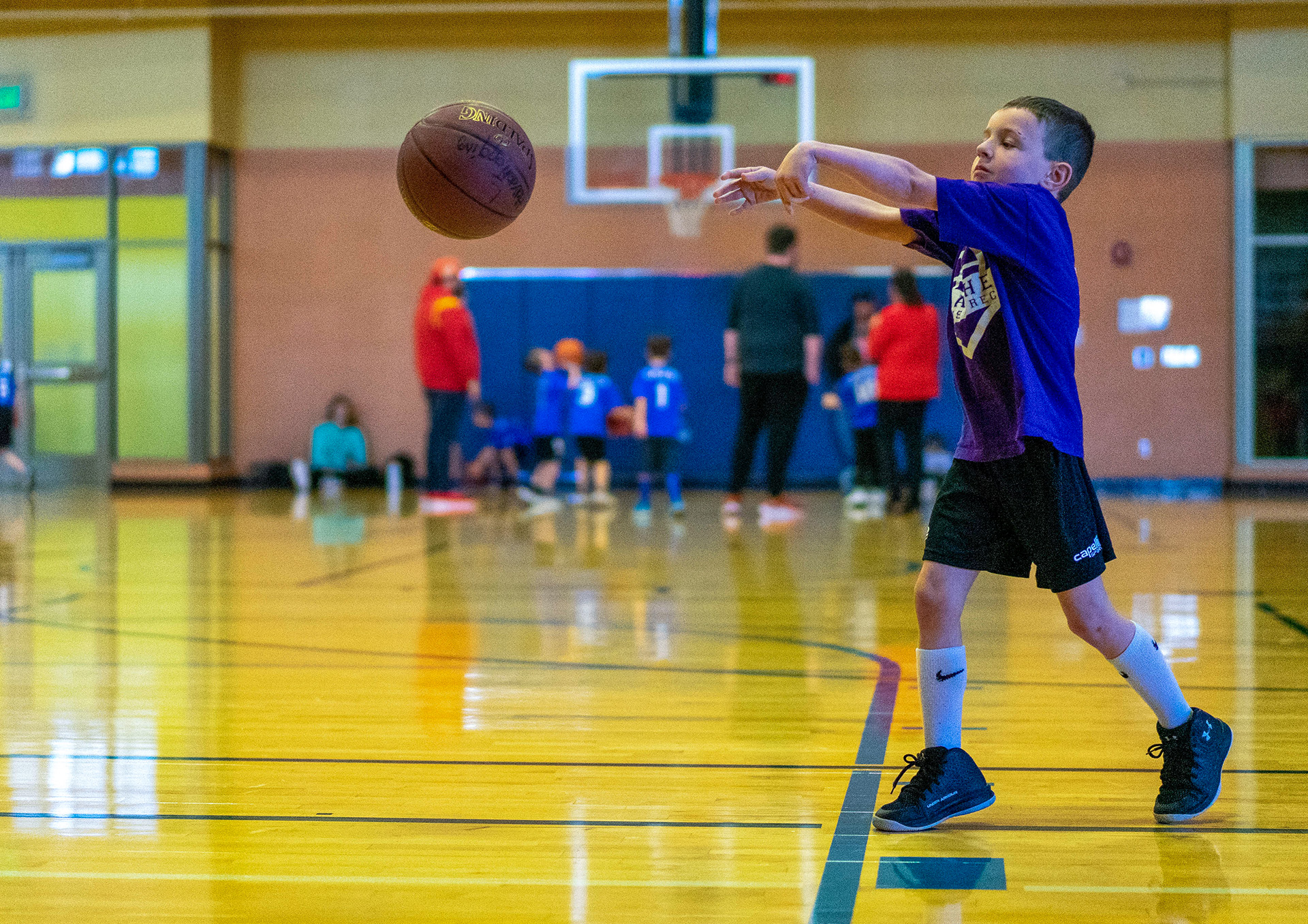 A child wearing a purple shirt, standing on an indoor basketball court, passing a basketball.