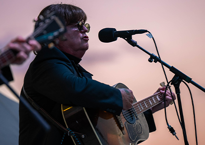 A man dressed at John Lennon plays an acoustic guitar with a sunset sky in the background