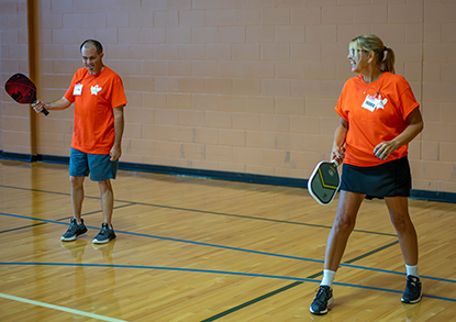 Two adults wearing orange shirts playing pickleball indoors