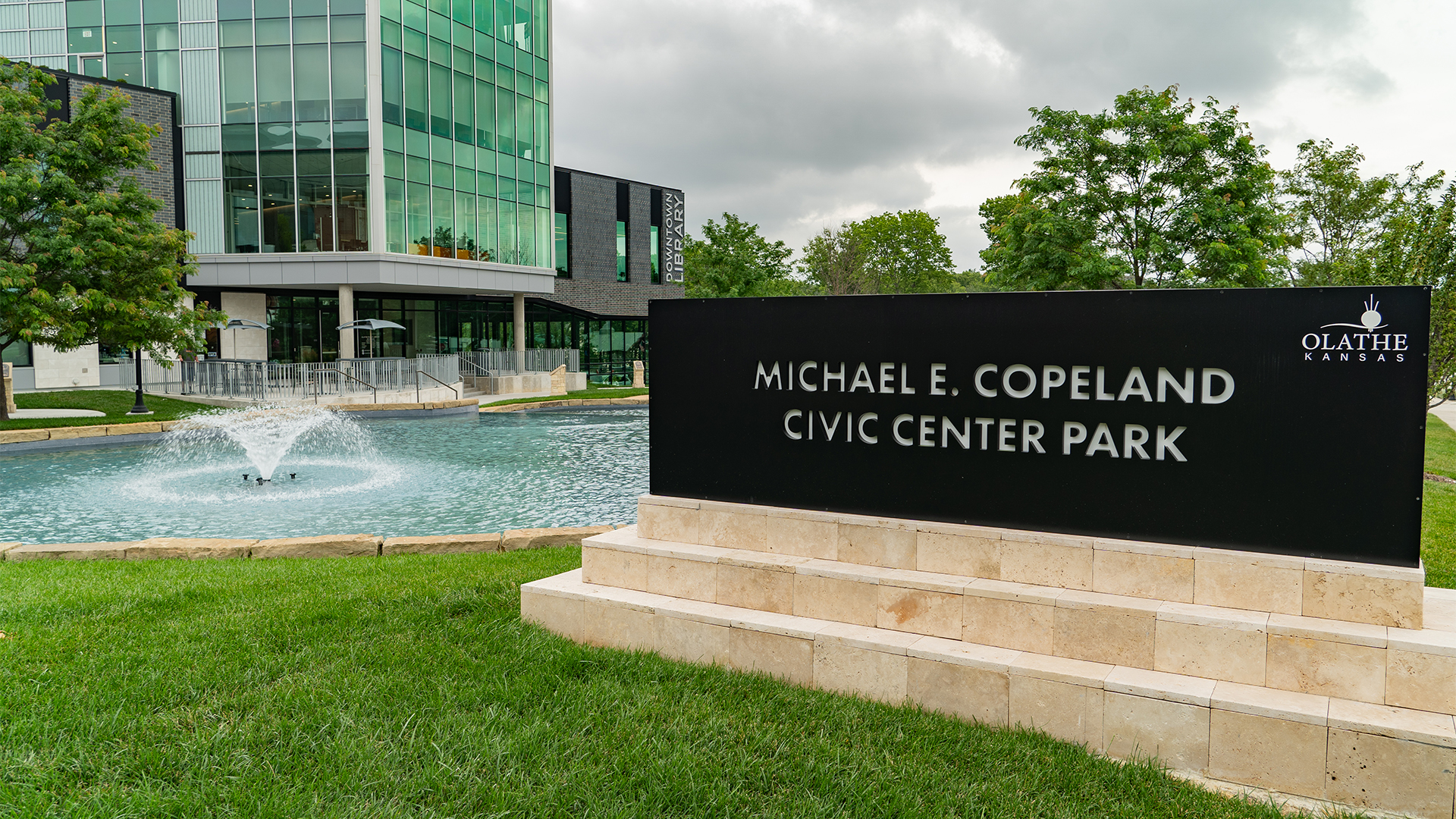 Sign stating "Michael E Copeland Civic Center Park" with fountain and Olathe Downtown Library in the background