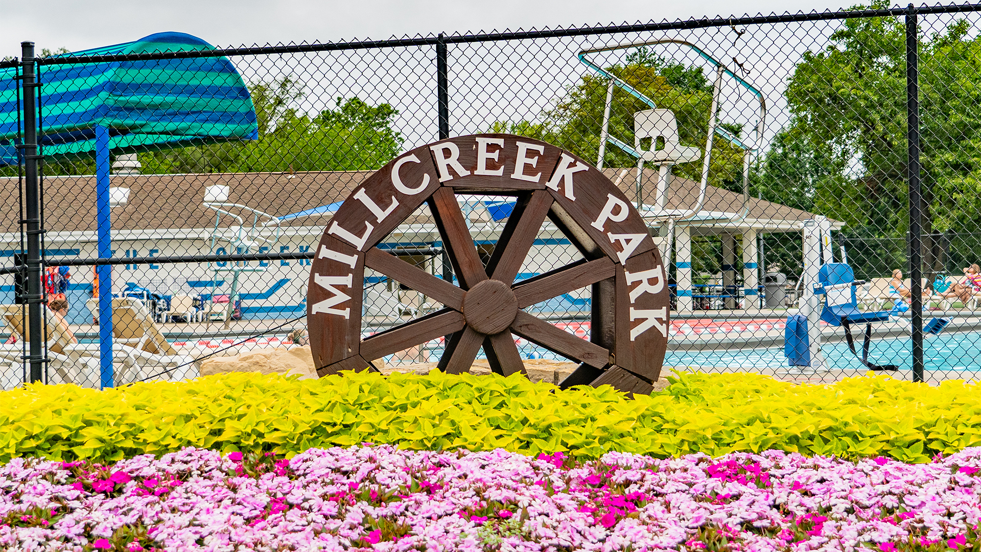 Wooden water wheel shaped sign stating "Mill Creek Park" in a bed of green plants and pink flowers with a pool in the background