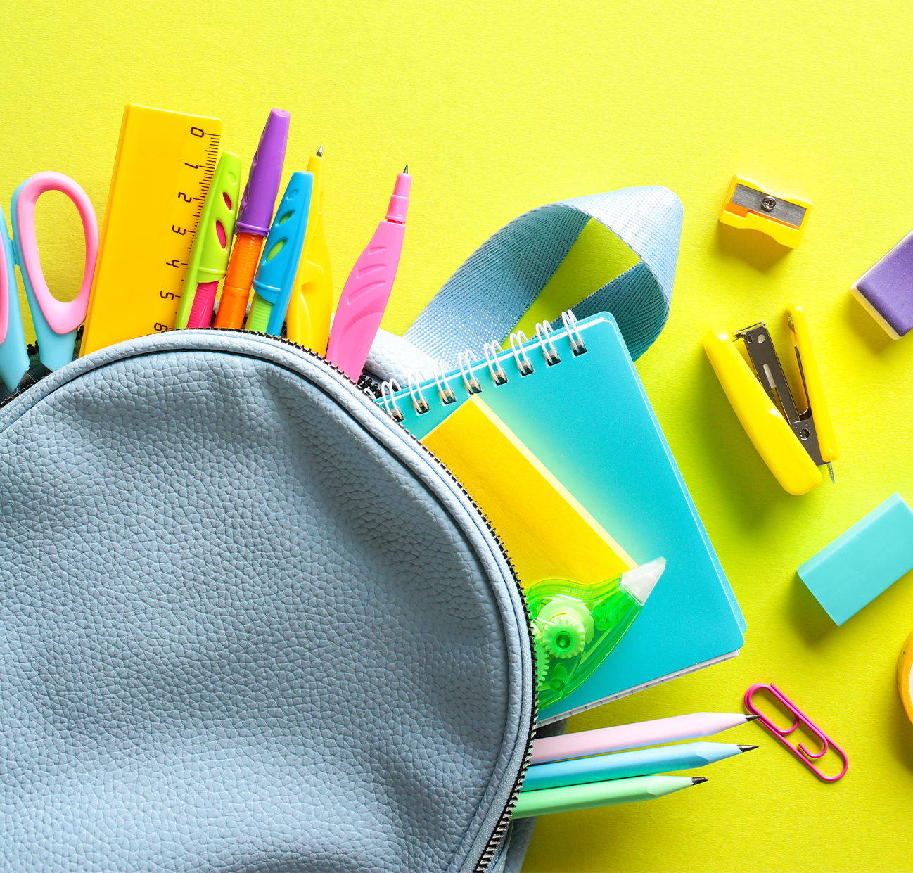 A light blue backpack with a variety of school supplies sticking out of it. The backpack and supplies are laying on a yellow backdrop.