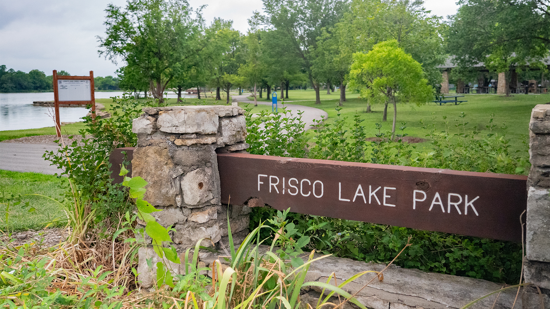 Wooden sign with stone pillars that states "Frisco Lake Park" with lake and park shelter in the background