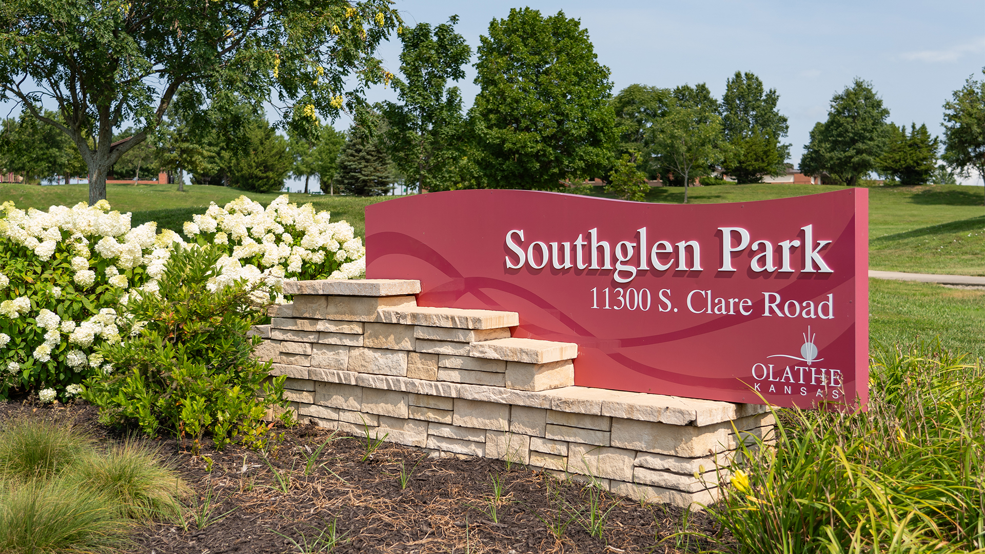 Red sign that states "Southglen Park" and "11300 S. Clare Road" with the City of Olathe logo, surrounded by a stone brick wall and white flowers