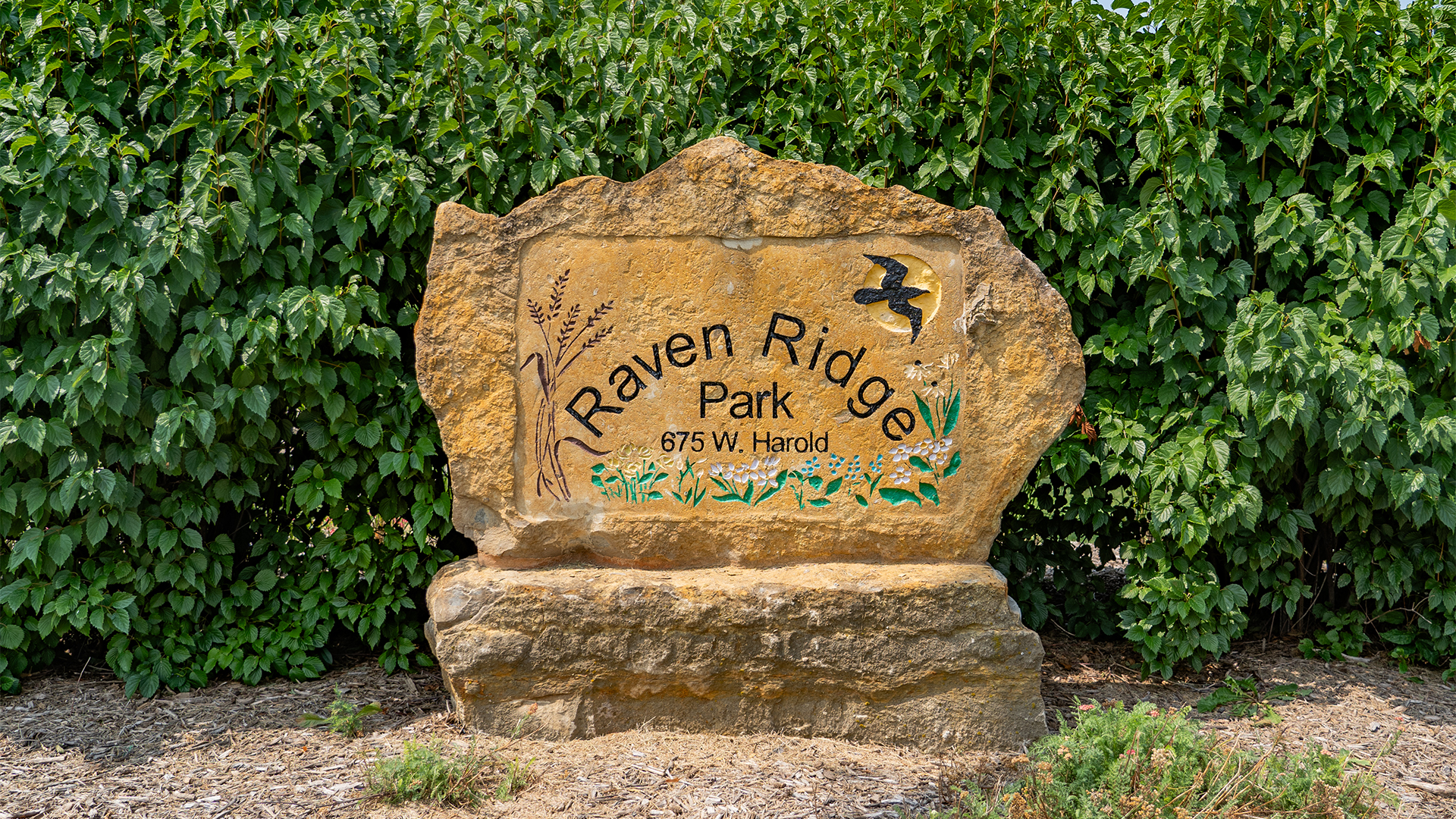 Stone sign that states "Raven Ridge Park" and "675 W. Harold" with paintings of wheat, flowers, a bird, and the sun