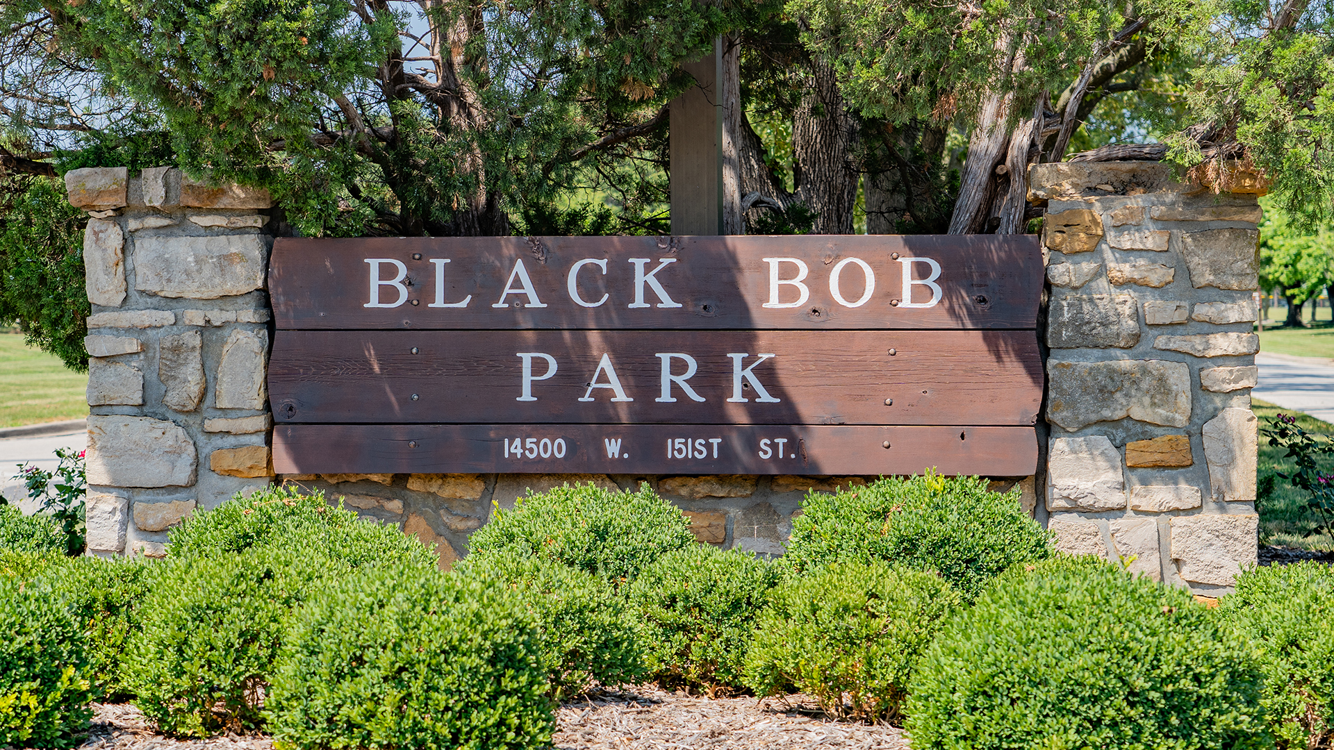 Wooden sign with stone brick pillars that reads "Black Bob Park" and "14500 W. 151st St." and bushes in the front