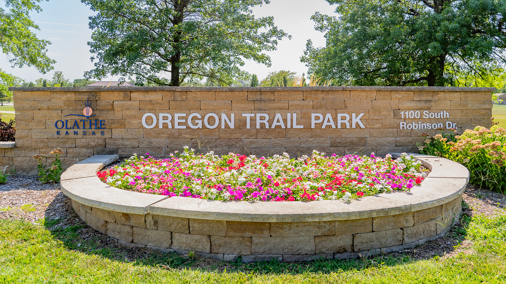 Stone brick sign that states "Oregon Trail Park" and "1100 South Robinson Dr." and has the City of Olathe logo and a bed of flowers in the front