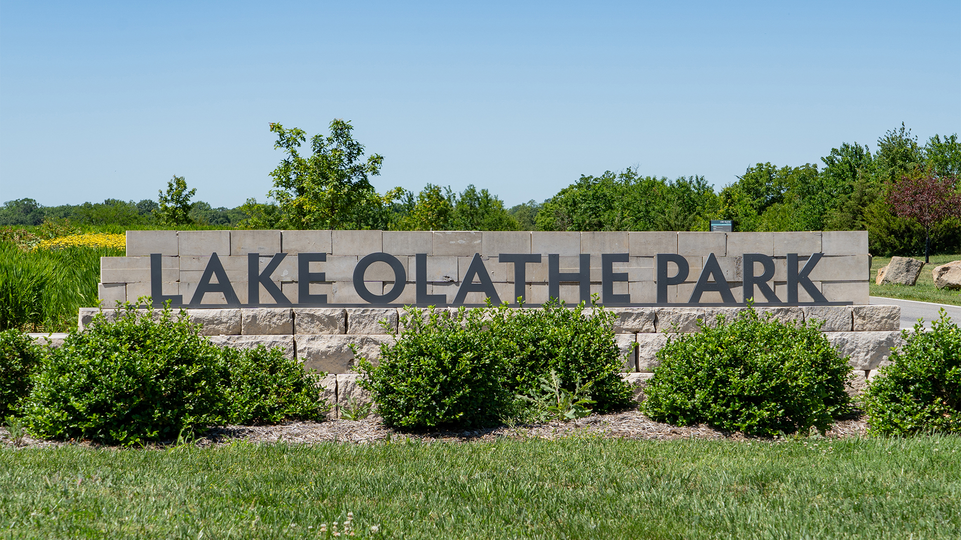 Stone brick sign that states "Lake Olathe Park" with bushes in the front