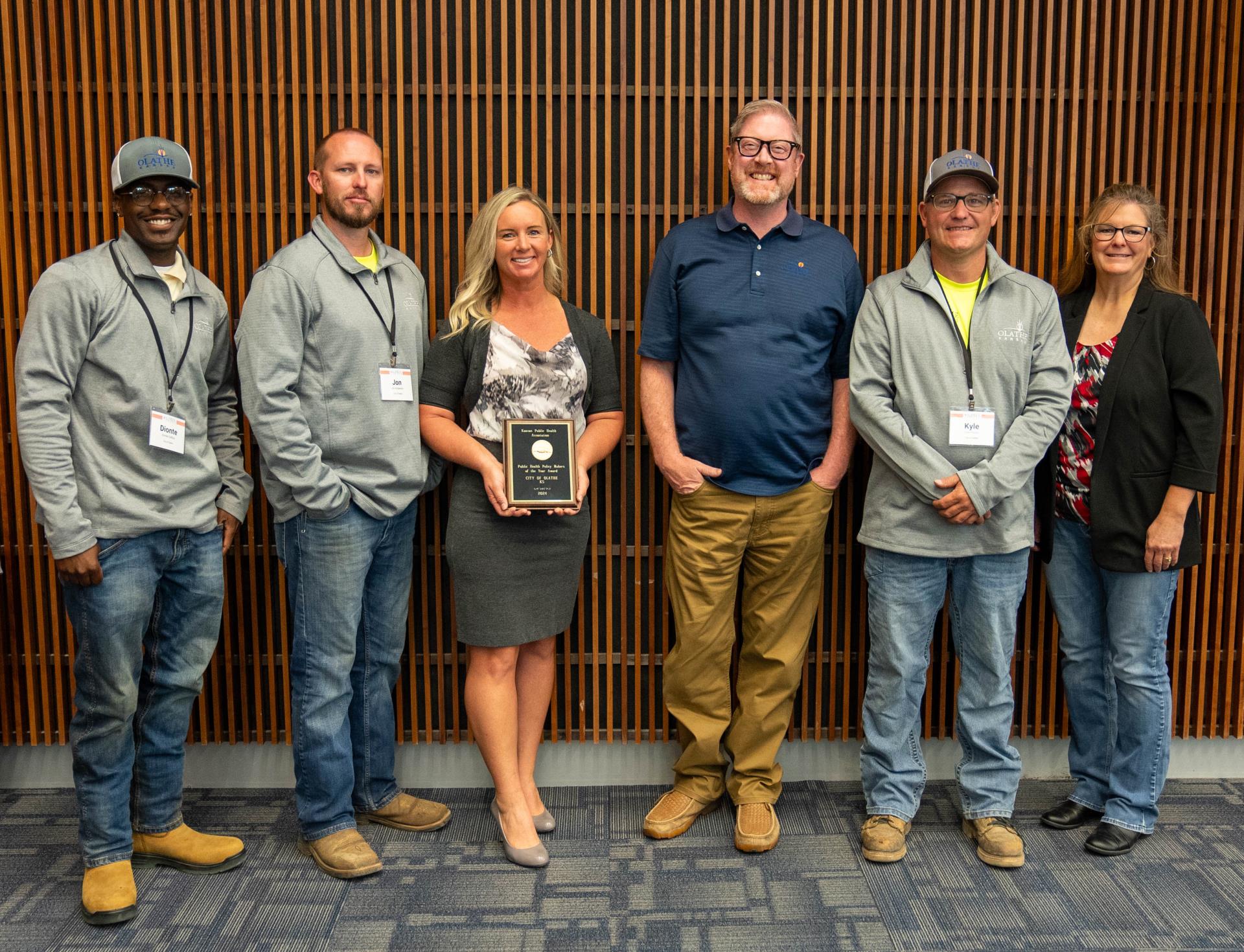 Six Olathe employees pose for a photo with an award, in front of a wood slat wall.